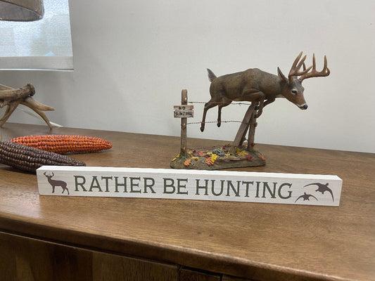 Decorative sign with 'Rather Be Hunting' text on a wooden surface with hunting-themed decor.