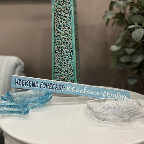 Decorative sign with 'Weekend Forecast' on a table with a decorative vase and plant in the background.