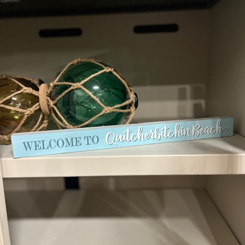 Decorative sign with 'Welcome to Quitcherlytchin Beach' on a shelf with decorative balls.