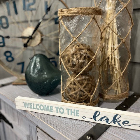 Decorative jars with twine, a clock, and 'Welcome to the Lake' sign on a wooden surface.