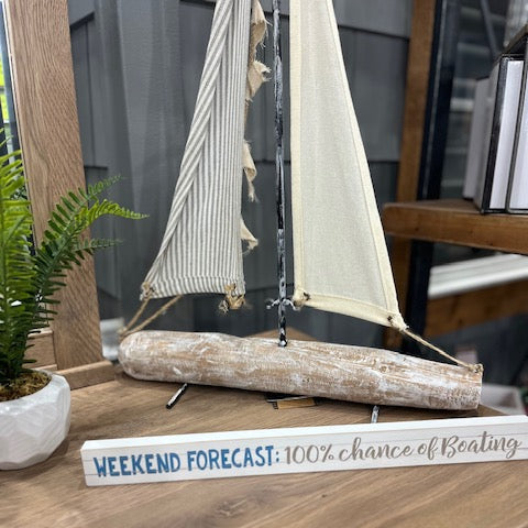 Decorative sailboat made of driftwood with striped sails on a wooden surface, next to a small sign with text.