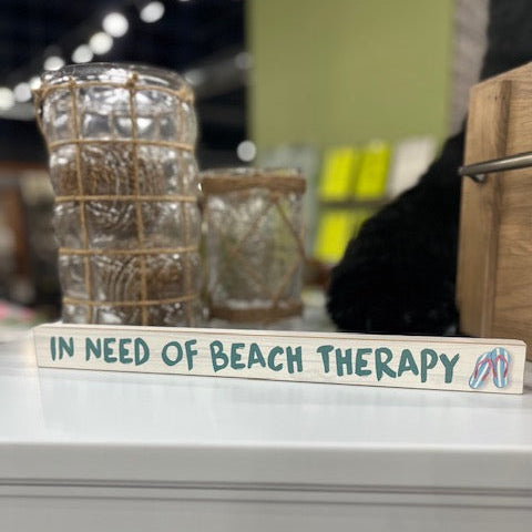 Decorative sign with 'In Need of Beach Therapy' text on a shelf.