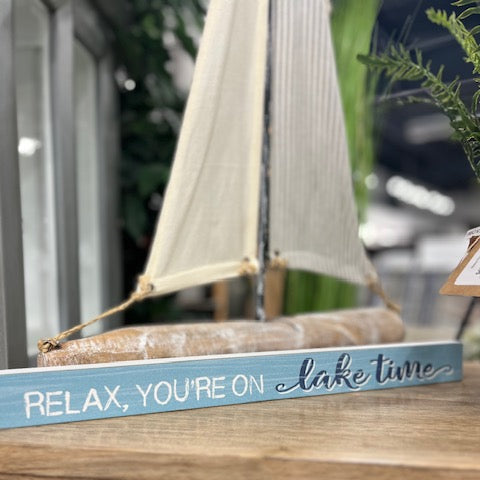 Decorative sign with sailboat design and 'Relax, you're on lake time' text on a wooden surface.