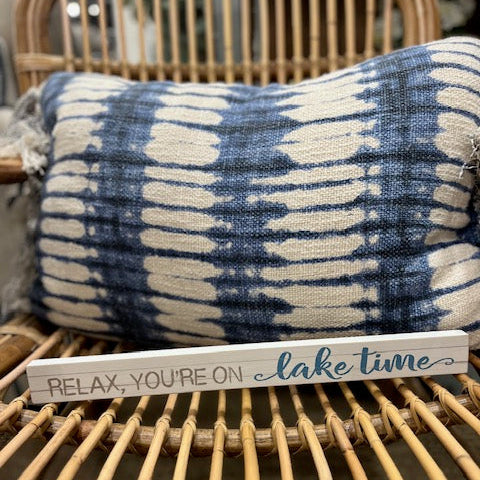 Decorative sign with 'Relax, you're on lake time' text on a wicker chair with a tie-dye pillow.