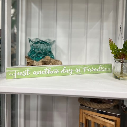 Decorative sign with 'just another day in Paradise' text on a shelf with plants and decor.