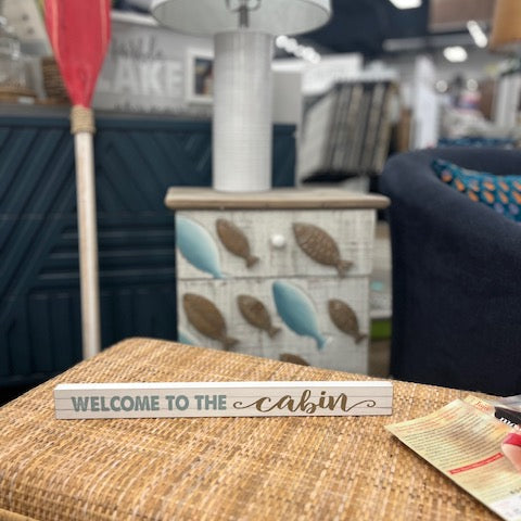 Decorative sign reading 'Welcome to the cabin' on a wicker surface with a blurred indoor background.