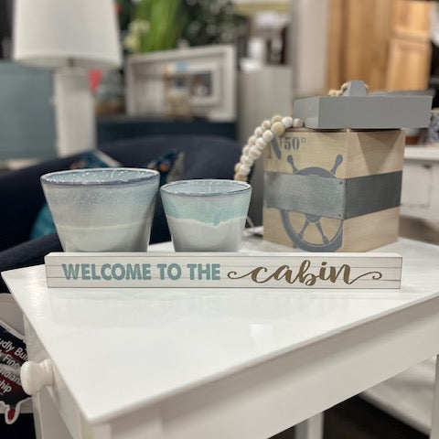 Decorative sign with 'Welcome to the cabin' on a white surface with two small pots.