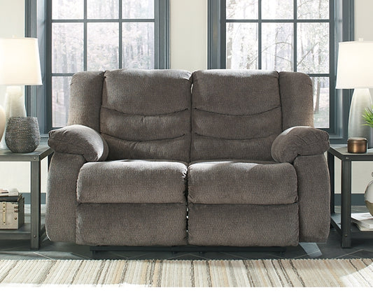 Gray reclining loveseat in a living room setting with windows and lamps.