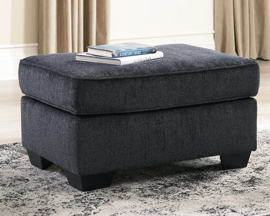 Gray ottoman with books on a rug in a room with curtains