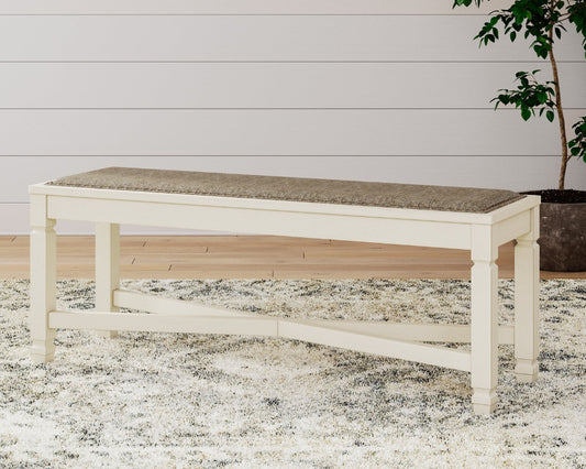 White bench with a textured cushion on a patterned rug, with a plant in the background.