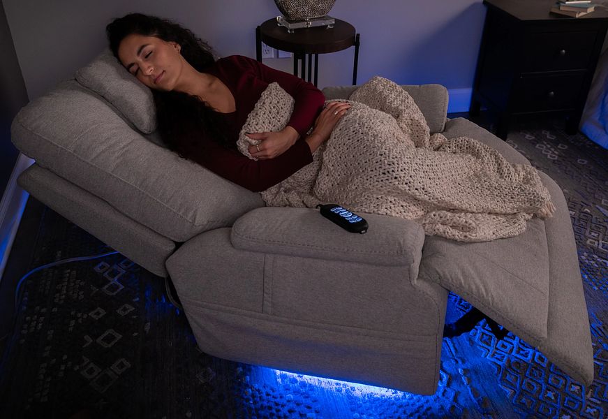 Woman lying on a recliner with a blanket, using a phone in a dimly lit room.