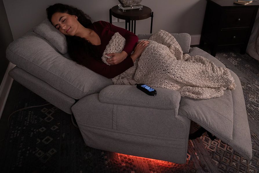 Woman lying on a gray recliner with a blanket, surrounded by dim lighting.