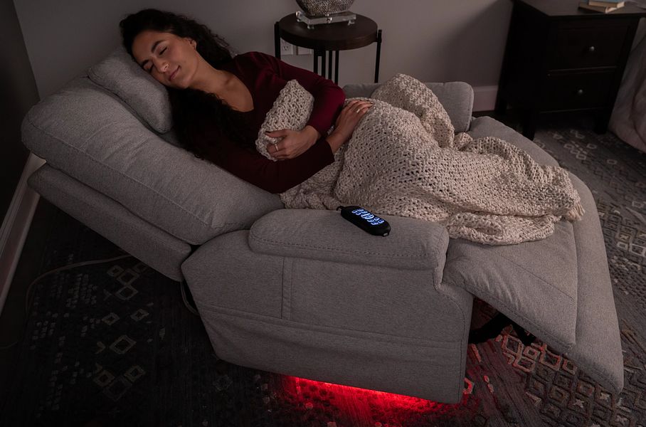 Woman relaxing on a gray recliner with a blanket, surrounded by dim lighting.