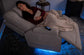Woman lying on a gray recliner with a blanket, surrounded by blue lighting.