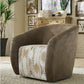 A modern barrel-style armchair in a styled living room. The chair features a taupe-brown suede-like fabric on the curved back and arms, with a distressed, rustic-patterned seat cushion in cream, beige, and brown tones.