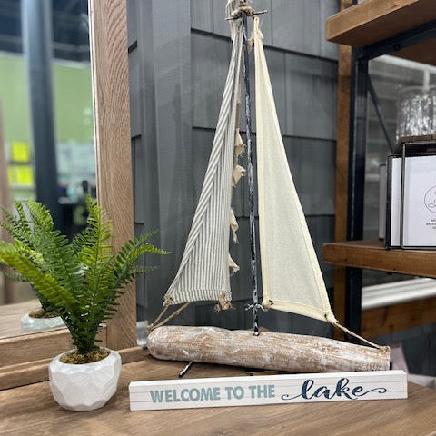 Sailboat model and 'Welcome to the Lake' sign on a wooden surface with a plant.