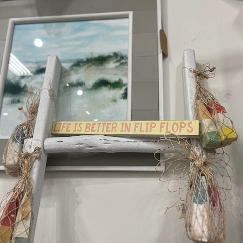 Decorative sign with 'Life is better in flip flops' on a white wall with a mirror and decorative elements.